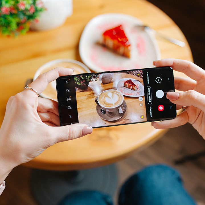 Χέρια κρατούν smartphone και φωτογραφίζουν λάτε με σχέδιο και φέτα τούρτας φράουλας σε ξύλινο τραπέζι.